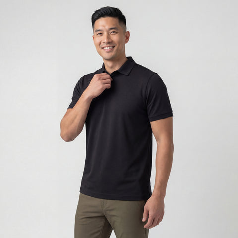 A man stands against a plain light background, smiling and wearing the Jack Archer Clubhouse Pique Polo in True Black and olive green pants, holding the collar of his moisture-managing shirt with one hand.