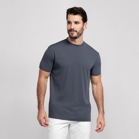 A man with short dark hair and a beard stands against a plain light gray background, wearing the Jack Archer Jetsetter Tee in Charcoal Gray and white pants. He looks slightly left and smiles softly.