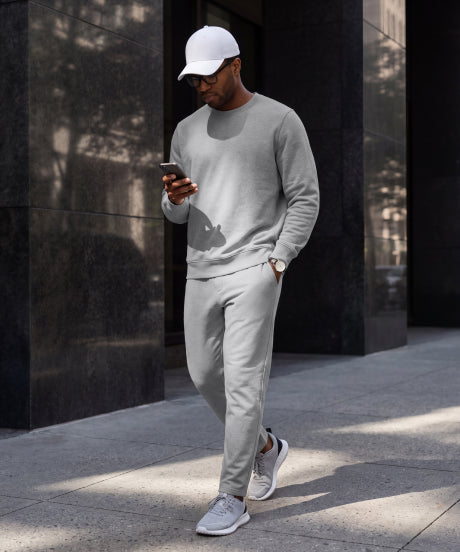 A man in a grey sweatshirt and joggers checking his phone on a city sidewalk.