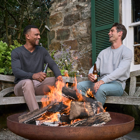 Two men in a charcoal V-neck sweater and a grey crewneck sitting by an outdoor fire pit with drinks.