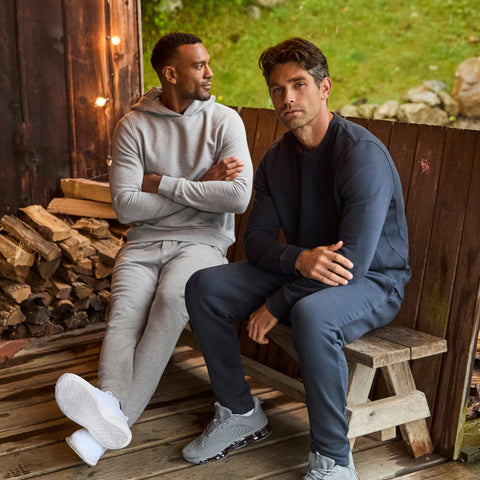 Two men in grey and navy fleece sweatsuits sitting on a cabin porch beside a stack of firewood.