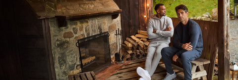 Two men in grey and navy fleece sweatsuits sitting on a cabin porch beside a stack of firewood.