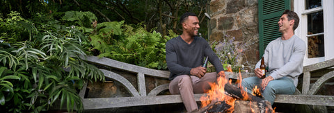 Two men in a charcoal V-neck sweater and a grey crewneck sitting by an outdoor fire pit with drinks.