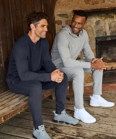 Two men in navy and grey fleece sweatsuits sitting on a cabin porch beside a stone fireplace.