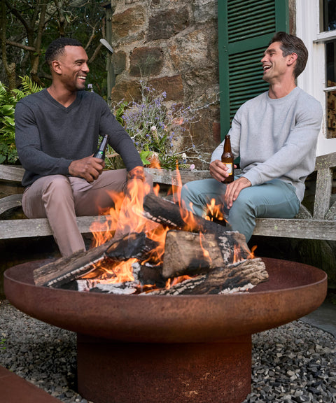 Two men in a charcoal V-neck sweater and a grey crewneck sitting by an outdoor fire pit with drinks.