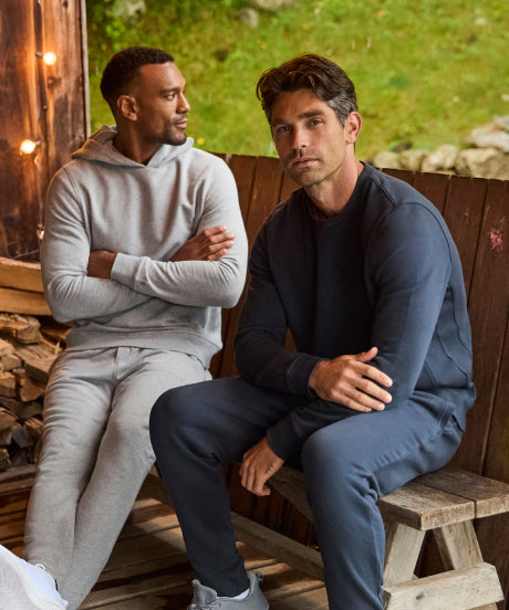 Two men in grey and navy fleece sweatsuits sitting on a cabin porch beside a stack of firewood.