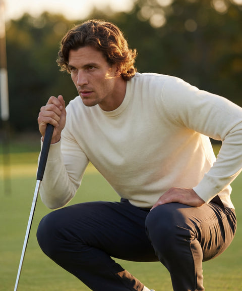 A man with wavy brown hair, in a cream sweater and Jack Archer's Jetsetter Tech Pant Slim Fit in Deep Blue, crouches on a golf course with a putter, focused on lining up his shot.