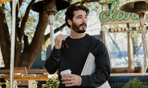 Man in Sandwash Crewneck in True Black holding a newspaper and coffee outside an oyster bar patio