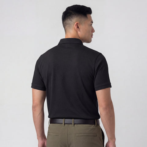 A man with short black hair is seen from behind, wearing the Jack Archer Clubhouse Pique Polo in True Black, paired with olive green pants and a black belt, standing against a plain light gray background.
