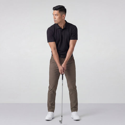 A man wearing the Jack Archer Clubhouse Pique Polo in True Black, brown pants, and white shoes stands indoors holding a golf club, poised to swing as he looks left against a plain light gray background.