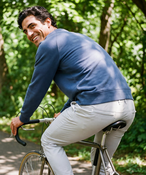 Man in blue crewneck sweater and Ice Gray Jetsetter Tech Pants riding a bike on a shaded path, smiling back