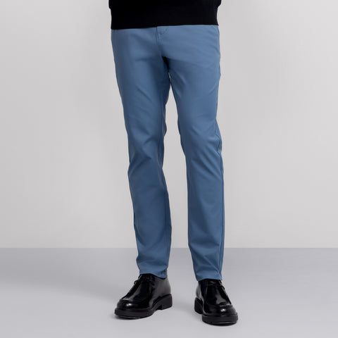 A person is shown from the waist down wearing Jetsetter Tech Pant Slim Fit in light blue and black dress shoes, standing against a plain, light background. The pants have a wrinkle-free finish and only the lower body is visible.
