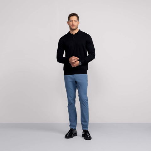 A man stands against a plain, light gray background wearing a black long-sleeve shirt, black shoes, and light blue Jetsetter Tech Pant Slim Fit, with his hands loosely clasped in front of him.