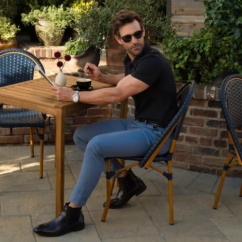A man in sunglasses, a black shirt, Jetsetter Tech Pant Slim Fit, and black boots sits at an outdoor café table with a coffee cup, a vase of flowers on the table, and plants in the background.