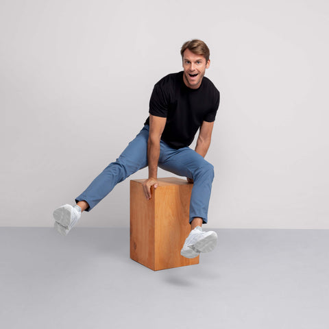 A man wearing the Jetsetter Tech Pant Slim Fit in blue and a black t-shirt sits playfully on a wooden box, balancing with his arms out and smiling. He pairs them with white sneakers against a plain, light background.
