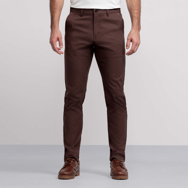 A man wears Jack Archer Jetsetter Tech Pant 3-Pack in brown, paired with a white shirt and brown leather shoes, standing against a light gray background. The image captures him from the chest down with his arms at his sides.