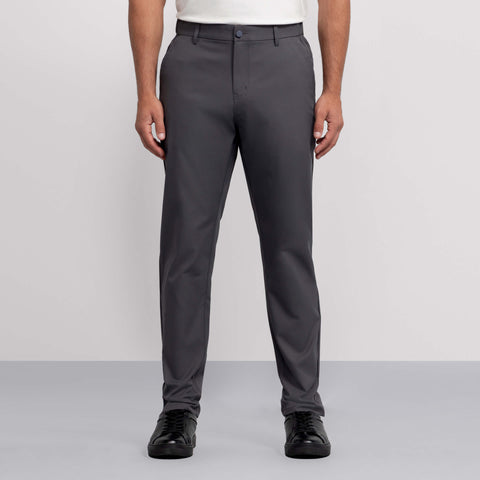 A person wears Jack Archer's Jetsetter Tech Pant in straight-fit charcoal gray, paired with a white tucked-in shirt and black shoes, standing against a plain light gray background. The image shows them cropped from the shoulders down.