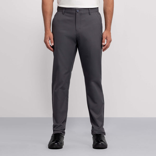 A person wears Jack Archer's Jetsetter Tech Pant in straight-fit charcoal gray, paired with a white tucked-in shirt and black shoes, standing against a plain light gray background. The image shows them cropped from the shoulders down.