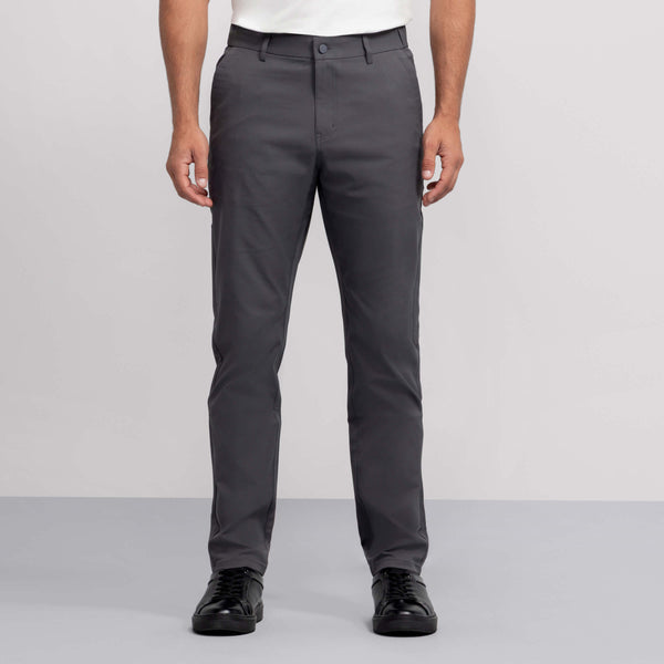 A person wearing Jetsetter Tech Pant Slim Fit in gray, paired with a white shirt and black sneakers, stands against a plain light gray background. The image displays the individual from shoulders to feet.