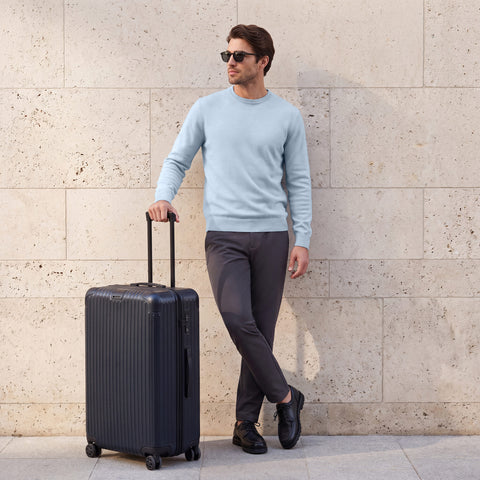 Charcoal Jetsetter pants and a light blue crewneck sweater, worn beside a rolling suitcase at a stone wall.