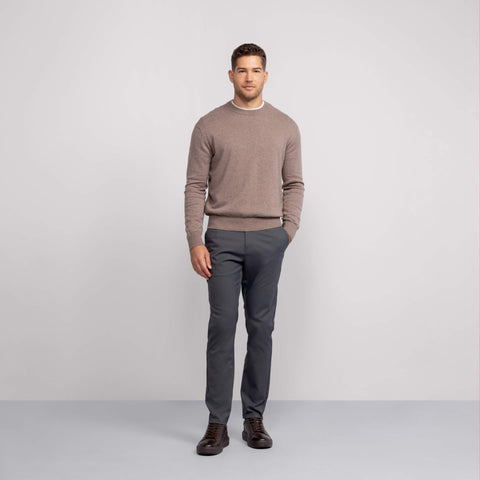 A man stands against a plain light gray background, wearing the Jetsetter Tech Pant Slim Fit in dark gray, paired with a light brown sweater and dark brown shoes. He has short hair, a neutral expression, and one hand in his pocket.