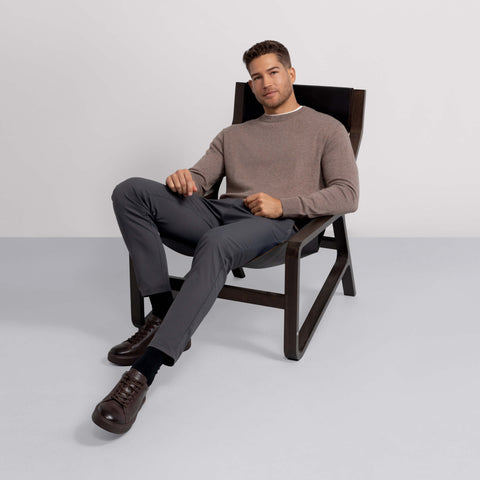 A man with short hair sits relaxed in a dark wooden chair, wearing a light brown long-sleeve shirt, Jetsetter Tech Pant Slim Fit, black socks, and dark brown shoes against a plain light background.