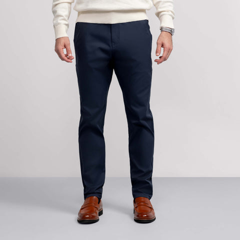 A man stands against a plain background, wearing Jack Archer's Jetsetter Tech Pant in navy—sleek, wrinkle-resistant—paired with a white sweater, brown dress shoes, and a watch. The image is cropped at the shoulders to show only his lower half.