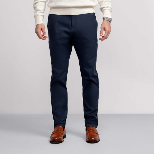 A man stands against a plain background, wearing Jack Archer's Jetsetter Tech Pant in navy—sleek, wrinkle-resistant—paired with a white sweater, brown dress shoes, and a watch. The image is cropped at the shoulders to show only his lower half.