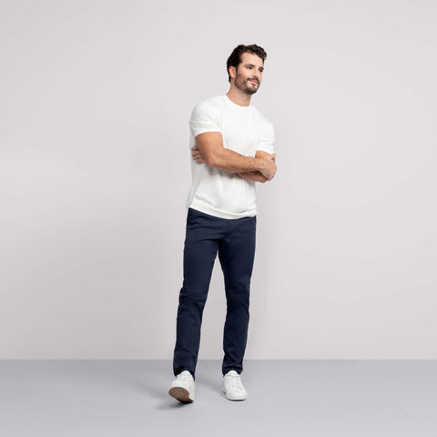 A man with short dark hair and a beard stands against a plain light background, arms crossed and relaxed, wearing a white T-shirt, white sneakers, and Jetsetter Tech Pant Straight Fit in dark blue.