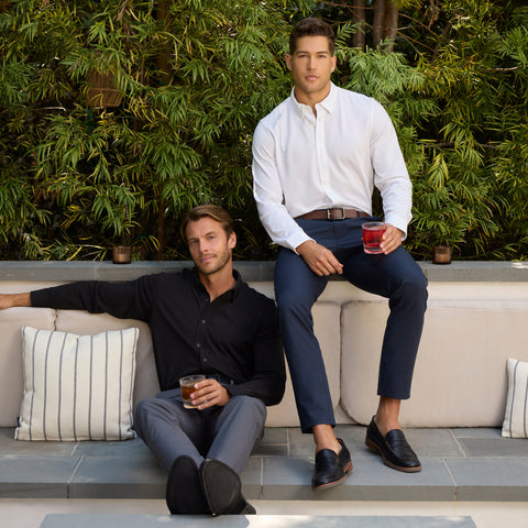 Two men relax with drinks on an outdoor sofa. One wears a black shirt and gray pants, and the other sports a white shirt and Jack Archer Jetsetter Tech Pant Slim Fit in Deep Blue. Greenery and striped pillows create a cozy background.