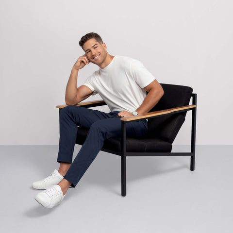 A man with short hair sits sideways in a modern black chair, smiling. He's wearing a white t-shirt, Jack Archer Jetsetter Tech Pant Slim Fit in Deep Blue, and white sneakers. One arm rests on the chair's wooden armrest against a plain background.