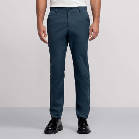 Against a plain background, a person wears Jack Archer's teal Jetsetter Tech Pant—water-repellent and wrinkle-resistant—paired with a white tucked-in shirt and black dress shoes. The photo is cropped from the shoulders down.