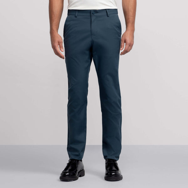 Against a plain background, a person wears Jack Archer's teal Jetsetter Tech Pant—water-repellent and wrinkle-resistant—paired with a white tucked-in shirt and black dress shoes. The photo is cropped from the shoulders down.