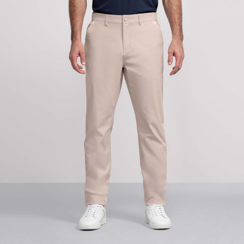 A man stands against a light gray background wearing Jack Archer’s Jetsetter Tech Pant in beige, paired with a tucked-in dark navy shirt and white sneakers.