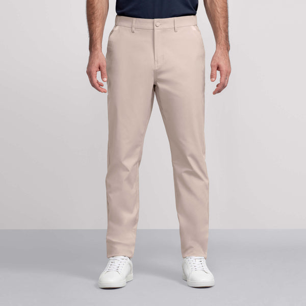 A man stands against a light gray background wearing Jack Archer’s Jetsetter Tech Pant in beige, paired with a tucked-in dark navy shirt and white sneakers.