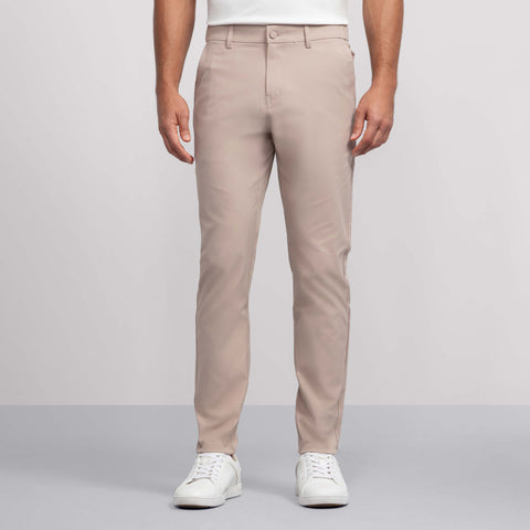 A person wears Jack Archer's Jetsetter Tech Pant Slim Fit in Oak Khaki with white sneakers, standing against a plain light gray background. Only their lower torso and legs are visible; the upper body is mostly out of frame.