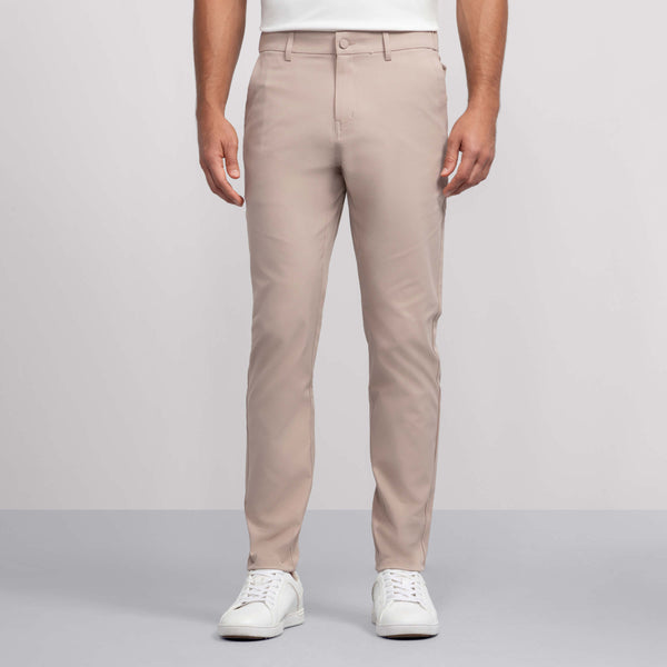 A person wears Jack Archer's Jetsetter Tech Pant Slim Fit in Oak Khaki with white sneakers, standing against a plain light gray background. Only their lower torso and legs are visible; the upper body is mostly out of frame.