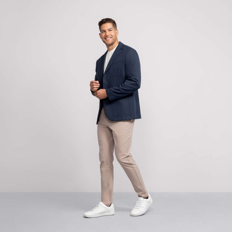 A man with short hair smiles against a light gray background, wearing a navy blazer, white tee, white sneakers, and Jack Archer Jetsetter Tech Pant Slim Fit in Oak Khaki. He poses with one hand near his chest and the other at his side.