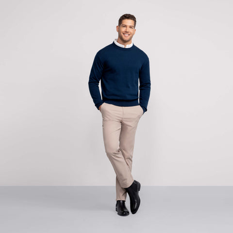 A man smiles with his hands in his pockets, wearing Jack Archer's Jetsetter Tech Pant Slim Fit in Oak Khaki, paired with a navy sweater, white collared shirt, and black dress shoes against a plain background.