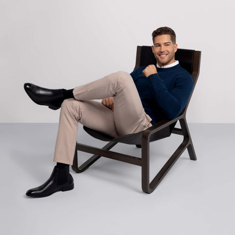 A man, dressed in Jack Archer's Jetsetter Tech Pant Slim Fit in Oak Khaki, pairs them with a navy sweater, white shirt, black socks, and dress boots. Relaxed in a modern chair, he smiles with one arm resting by his chin.