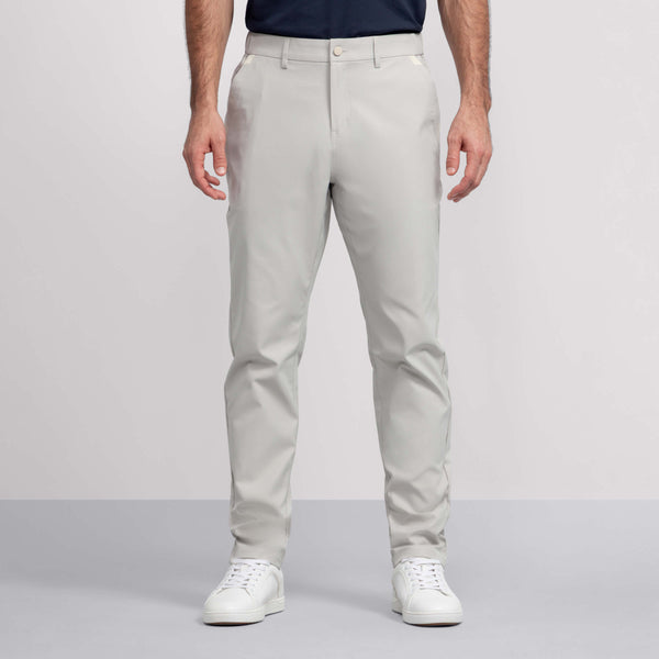A man wears Jack Archer Jetsetter Tech Pant Relaxed Fit in Sand Stone with a dark tucked-in shirt and white sneakers, standing with arms at his sides against a plain background. Only his upper body is partially visible.