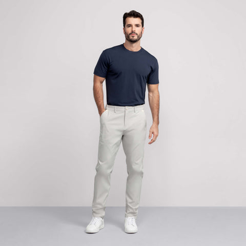 A man stands against a plain light background wearing a navy blue t-shirt, Jack Archer Jetsetter Tech Pant Relaxed Fit in Sand Stone, and white sneakers. He has one hand in his pocket and a neutral expression.