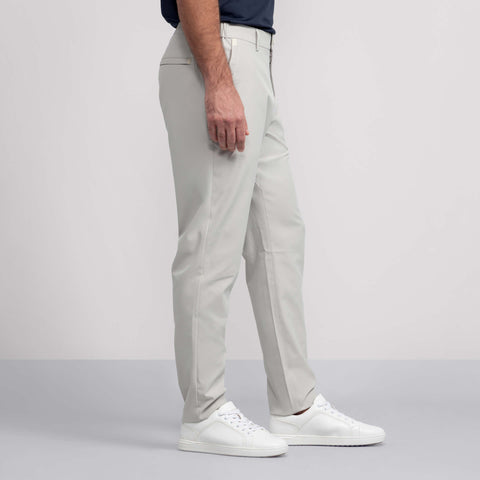 A man wearing Jack Archer's Jetsetter Tech Pant Relaxed Fit in Sand Stone, paired with a navy shirt and white sneakers, stands sideways against a plain light background.