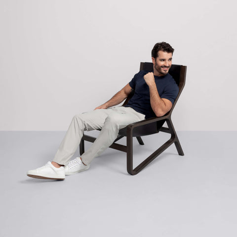 A man in a navy shirt, Jack Archer Jetsetter Tech Pant Relaxed Fit in Sand Stone, and white sneakers sits smiling in a modern wooden lounge chair against a plain, light background.
