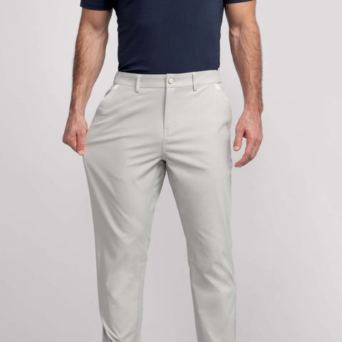 A man stands against a plain light background, arms relaxed at his sides, wearing a navy blue short-sleeve polo and Jack Archer Jetsetter Tech Pant Relaxed Fit in Sand Stone.