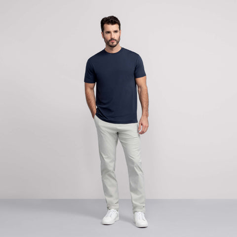 A man with short dark hair and a beard stands against a light gray background, wearing Jack Archer’s Jetsetter Tech Pant Straight Fit in Sand Stone, a navy blue t-shirt, and white sneakers. One hand rests in his pocket as he looks slightly to the side.