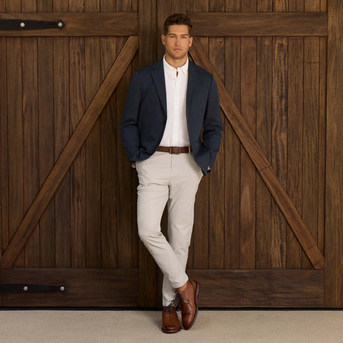 A man with short hair stands before large wooden doors, wearing a navy blazer, white shirt, and Jack Archer Jetsetter Tech Pant Straight Fit in Sand Stone, paired with brown dress shoes and a matching belt. His hands are in his pockets.