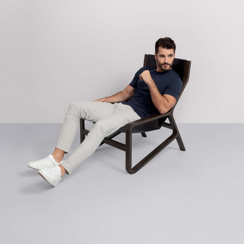 A man in a navy t-shirt, Jack Archer’s Jetsetter Tech Pant Straight Fit in Sand Stone, and white sneakers sits sideways on a modern black lounge chair, looking off to the side in a minimal, light-colored room.