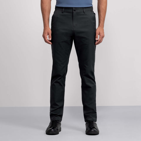 Wearing Jack Archer's Jetsetter Tech Pant—a dark, water-repellent, wrinkle-resistant pair—plus a blue short-sleeve shirt and black sneakers, a person stands against a plain light gray background, shown from shoulders to feet.