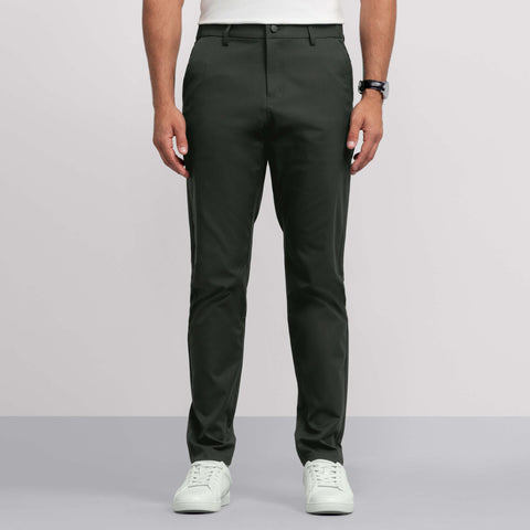A person wears dark green Jack Archer Jetsetter Tech Pant—wrinkle-resistant and water-repellent—with a white shirt, black wristwatch, and white sneakers, standing against a plain light background; only the lower half is visible.